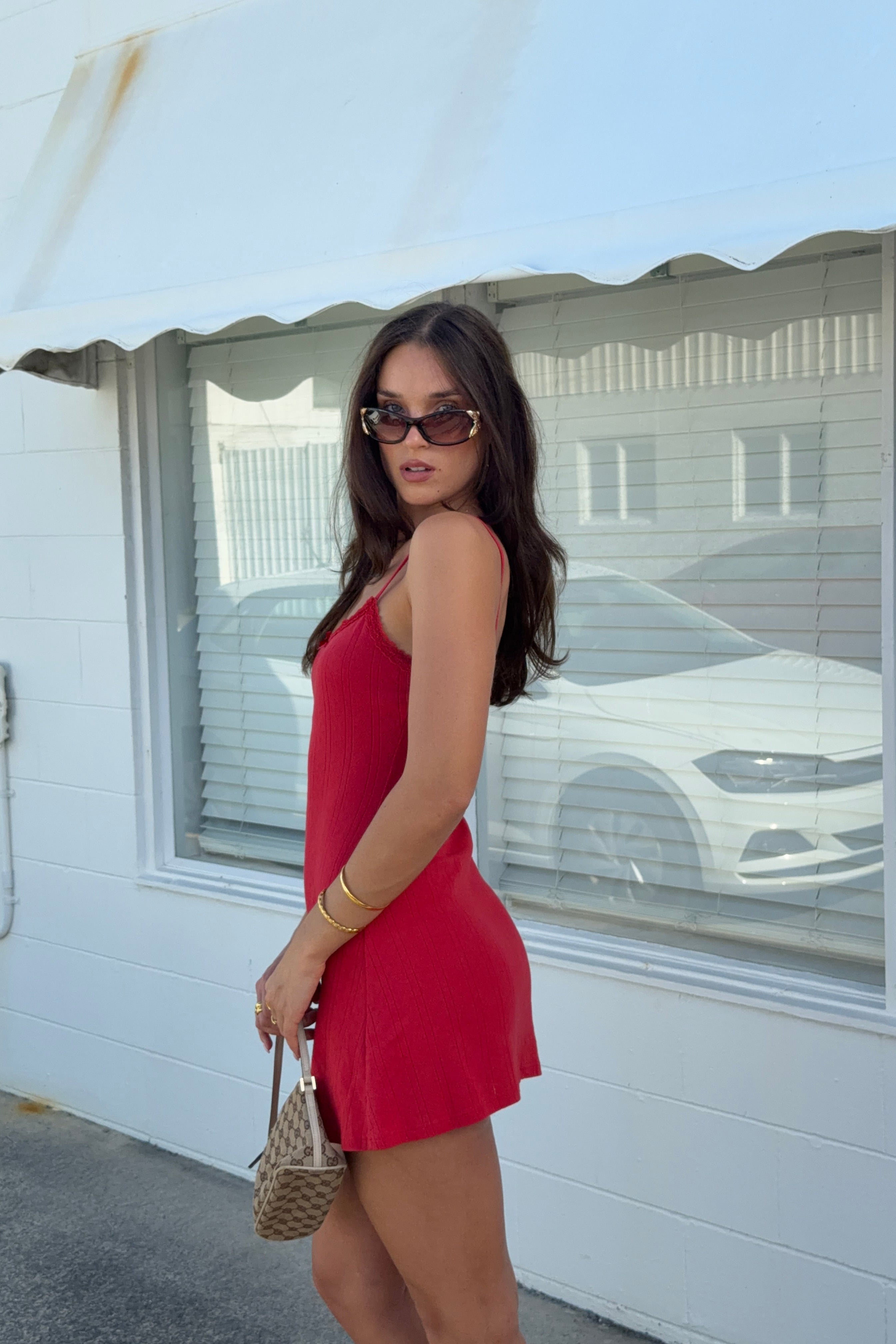 A woman wearing a bright red sleeveless mini dress and sunglasses stands outdoors in front of a white building with a window, holding a small woven handbag.