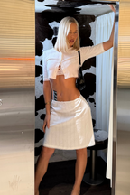 A woman with platinum blonde hair models a white ribbed crop top and matching white ribbed mini skirt set inside a photobooth, framed by a large metallic mirror. 