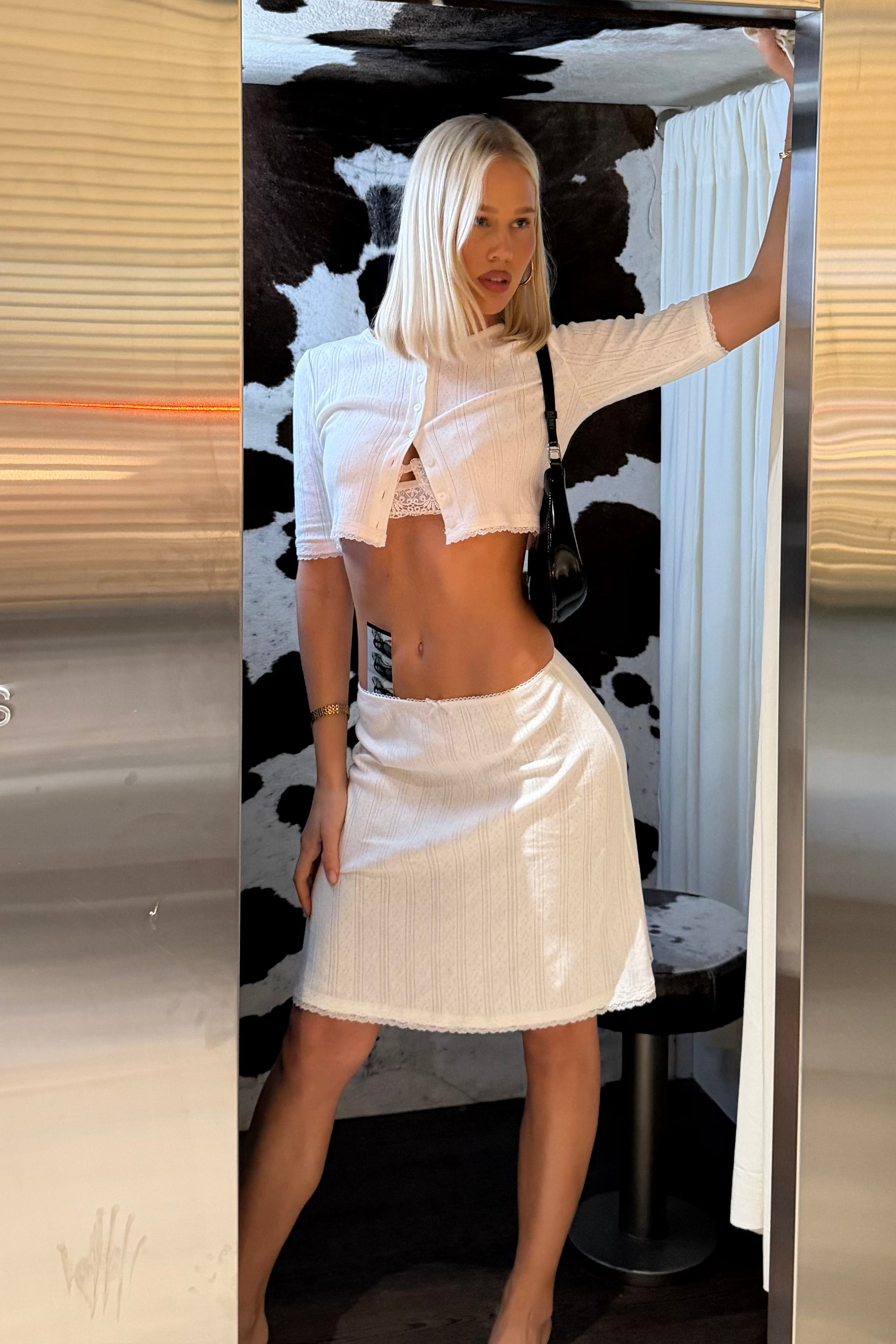 A woman with platinum blonde hair models a white ribbed crop top and matching white ribbed mini skirt set inside a photobooth, framed by a large metallic mirror. 