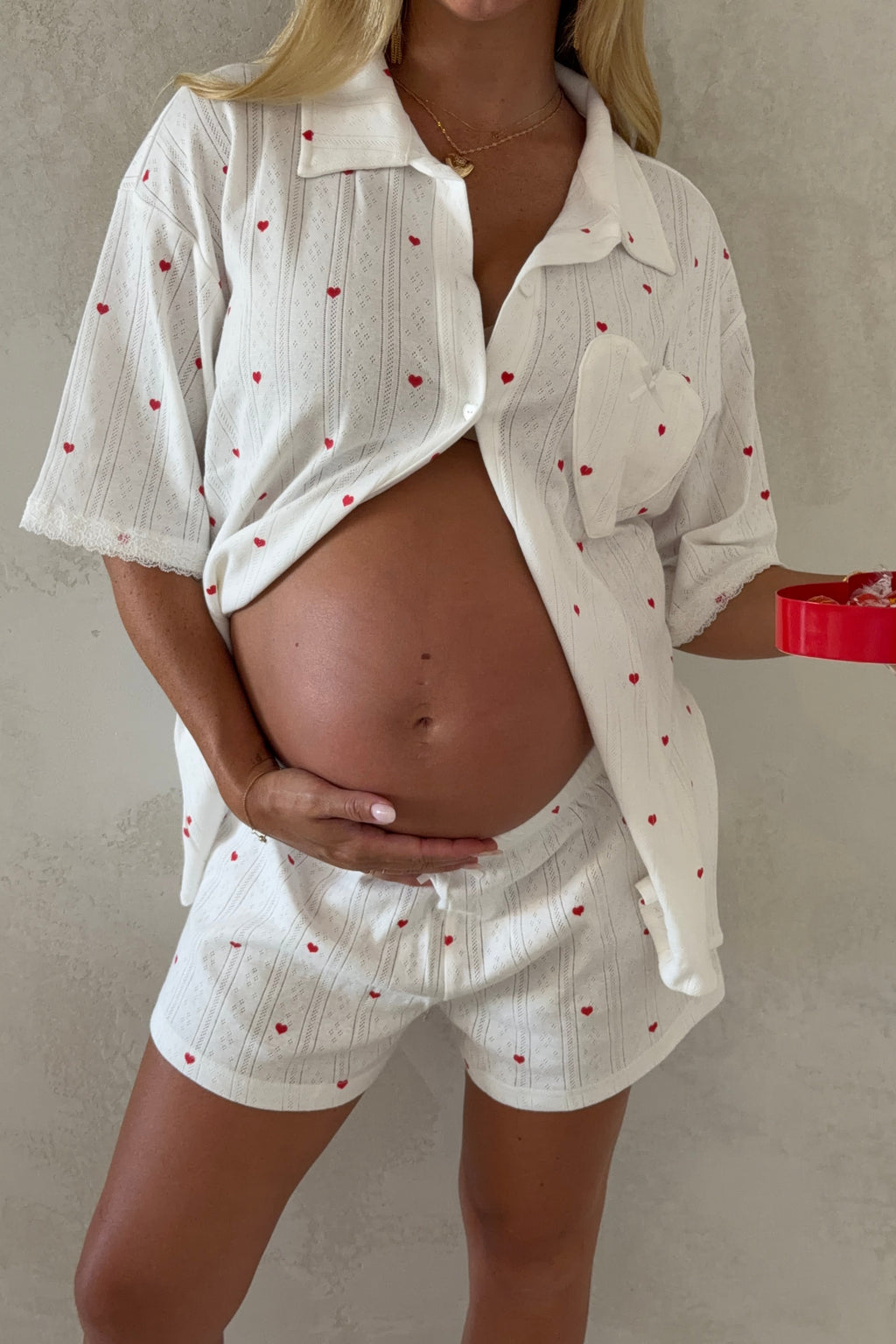 A pregnant woman wearing a white textured cotton pajama set with small red heart prints, consisting of a short-sleeve button-up shirt and matching shorts. She is holding a red heart-shaped gift box against a neutral textured wall.