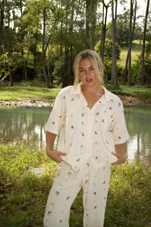 A person wearing a bunny-themed sleepwear set with a button-up top and long pants, featuring a heart-shaped pocket and lace trim, standing near a body of water.
