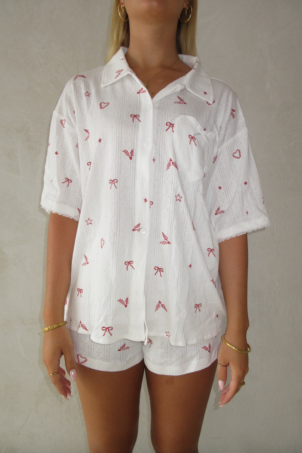 White christmas pajamas with red pattern worn by a girl on a plain background. Button up top with heart pocket, white heart buttons and lace trim on the sleeves and wide leg shorts 