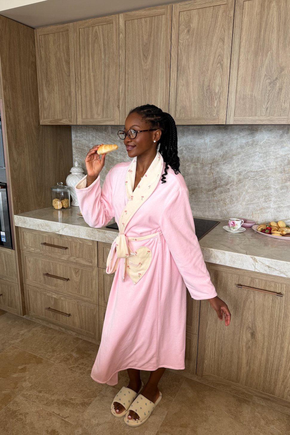 Woman in a pink robe standing in a kitchen eating a pastry.