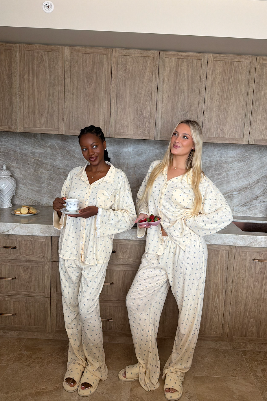 Two women wearing matching cream pajama sets with small blue floral prints stand in a kitchen setting. The set includes a long-sleeve top and matching pants, with one woman holding a teacup and saucer and the other holding strawberries.